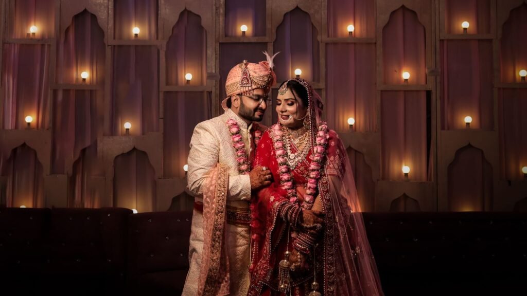 Indian newlywed couple in traditional clothing, smiling and holding hands indoors.