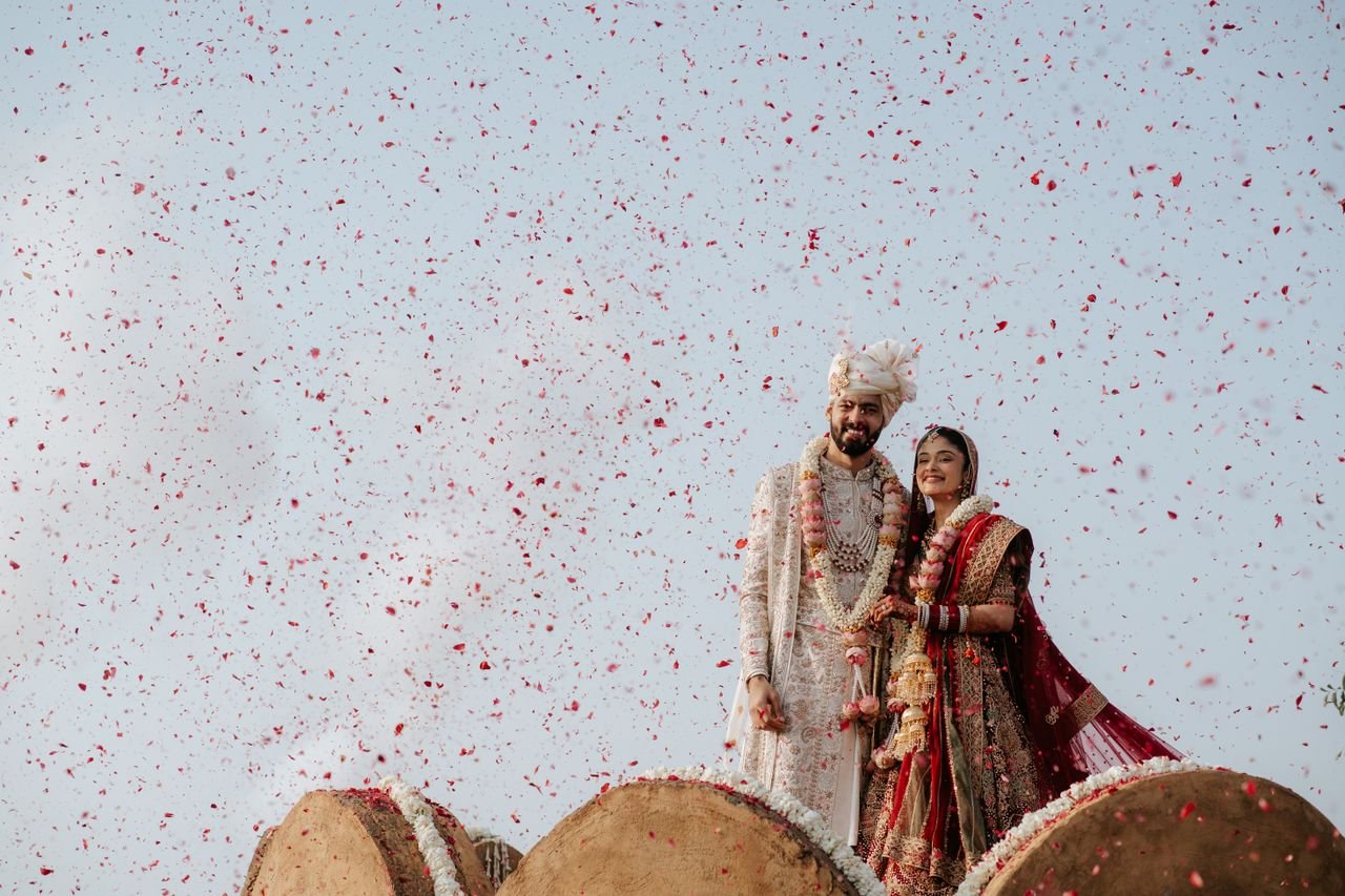 Beautiful Indian wedding couple adorned in traditional attire, celebrating under a shower of rose petals outdoors.